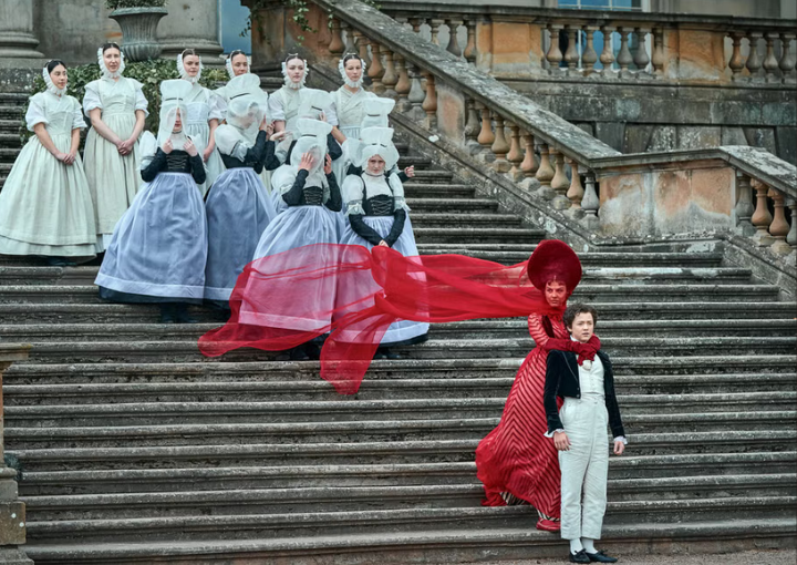 Жена в червени дрехи стои права на стълби на имение, прегърнала е момче.